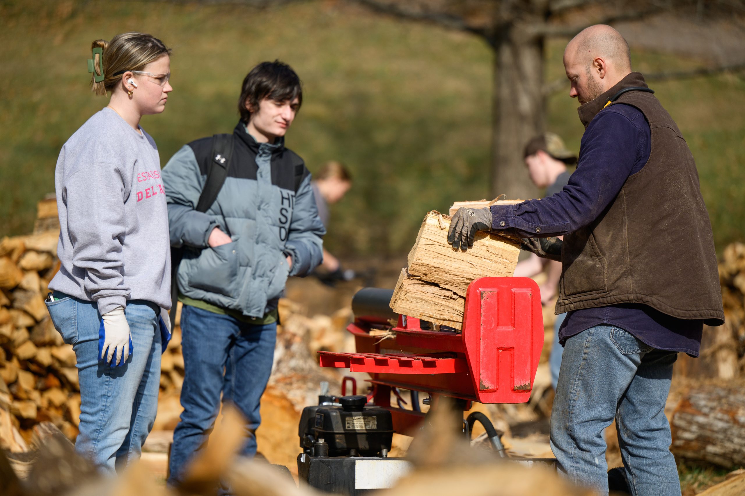 Man with beard demonstrates wood splitter to two young people outdoors in yard